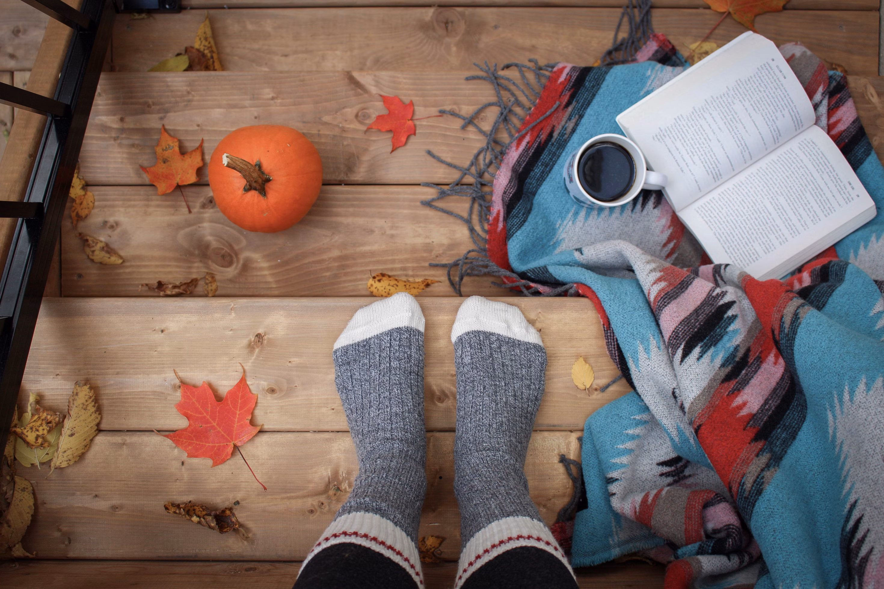 A POV shot of a pumpkin, blanket, drink, book and a female wearing socks on wooden decking.