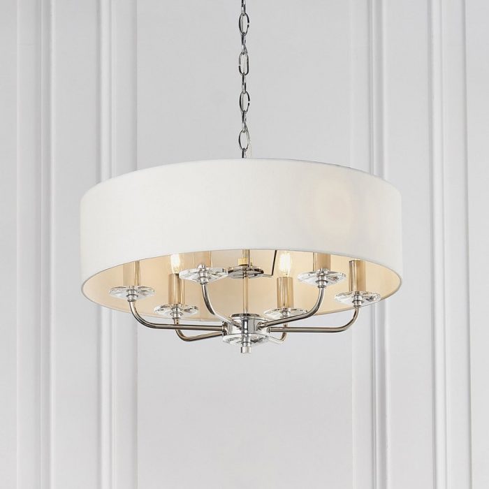 A pendant light with a nickle finish hanging from the ceiling of a white-walled room. The lamp shade is white/cream and the fixture replicates a candelabra.